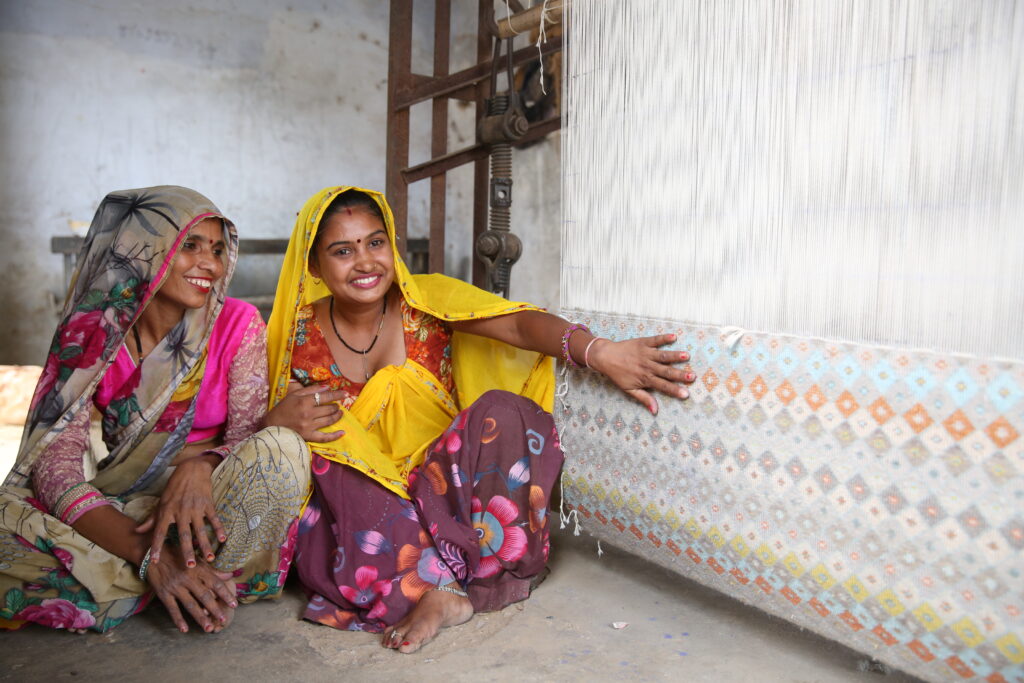 Two women in saris sit next to a rug loom