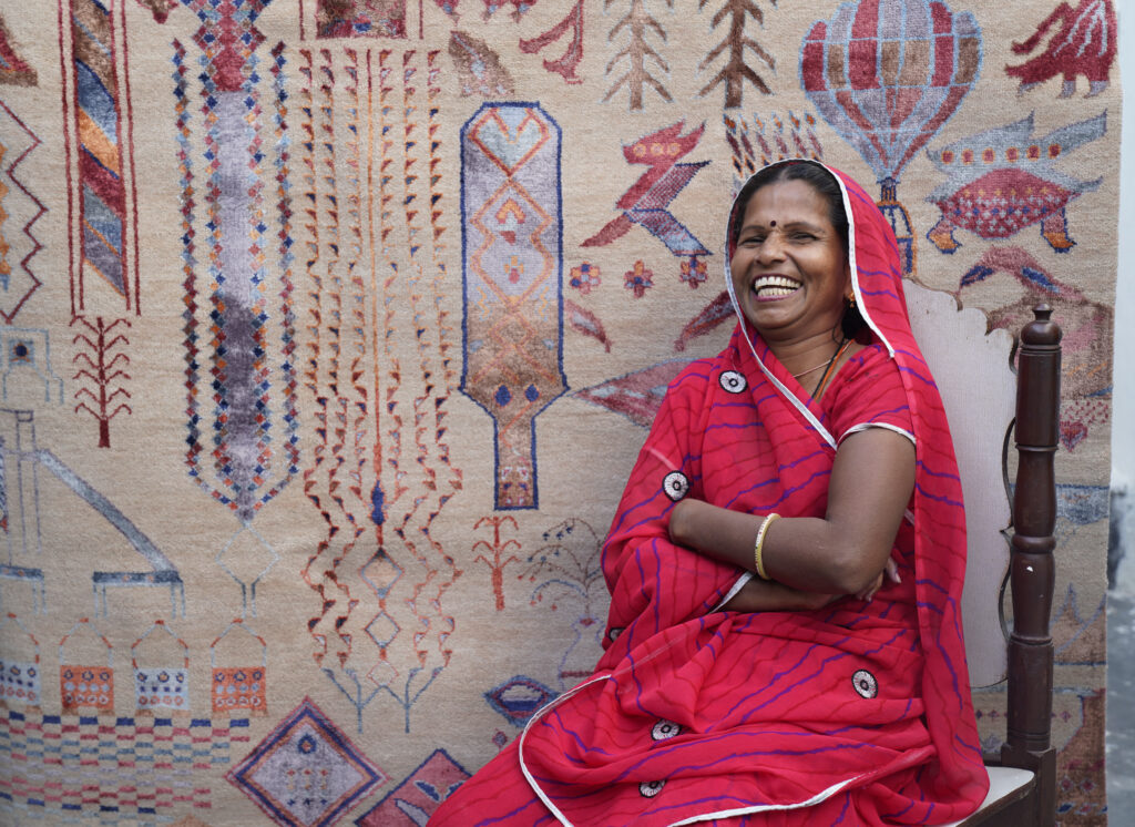 A woman in a bright orange sari sits in front on the rug she made
