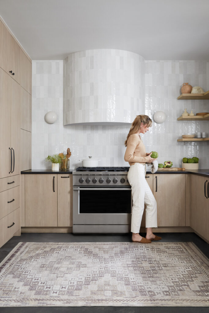 A woman stands in a neutral colored kitchen with a tan and beige pattern rug underfoot.