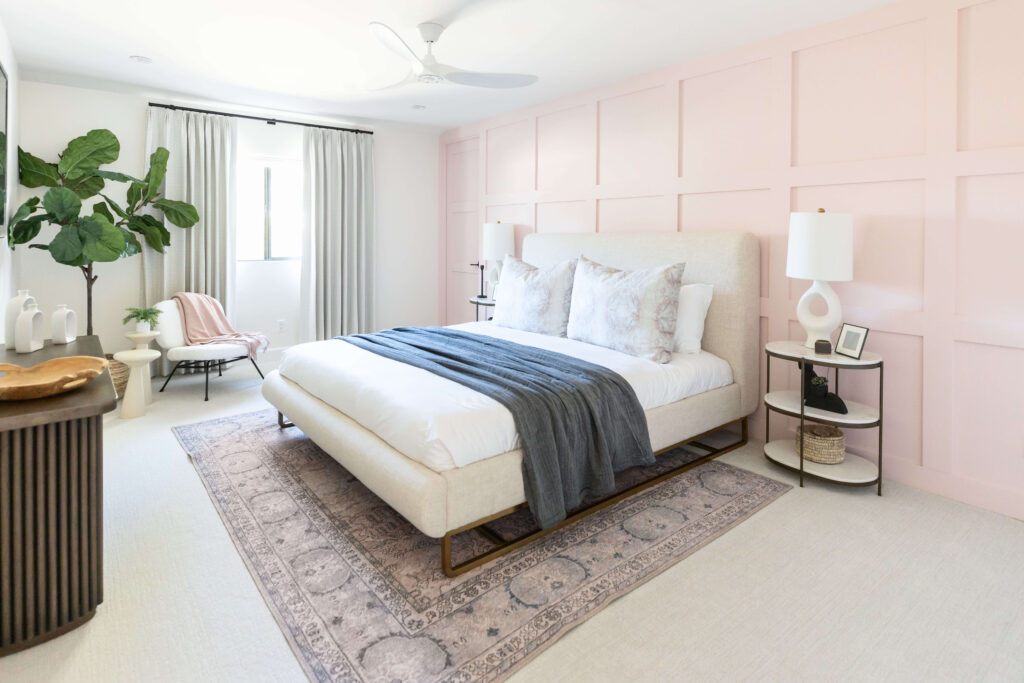 A bedroom with light pink accent wall, white bed, and pink and blue patterned rug