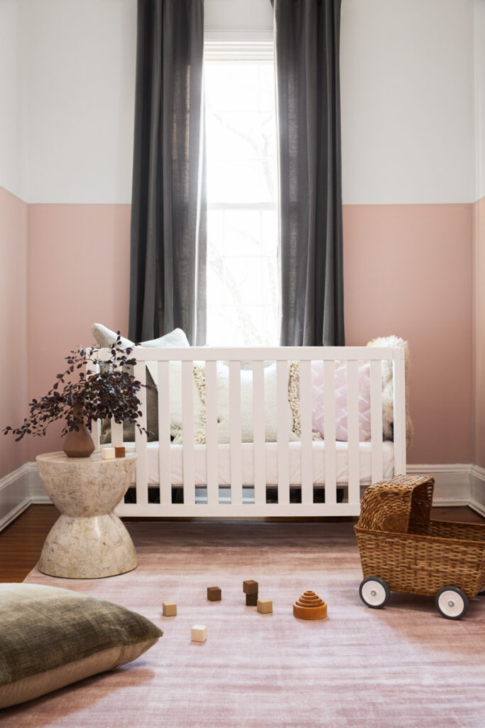 A nursery with white crib, dark gray drapes, pink walls, and pink rug