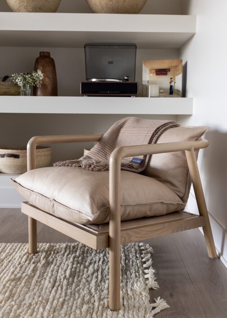 A light brown leather chair with wood arms sits in front of a bookcase and on top of a cream boucle style rug