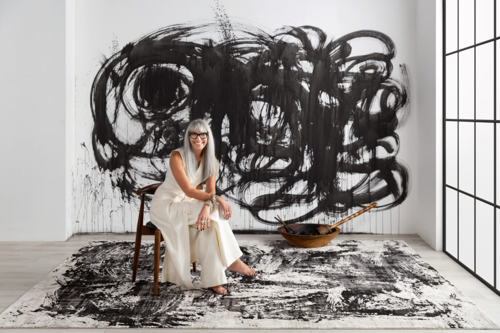 A woman in an all white jumpsuit sits in front of a black and white abstract mural