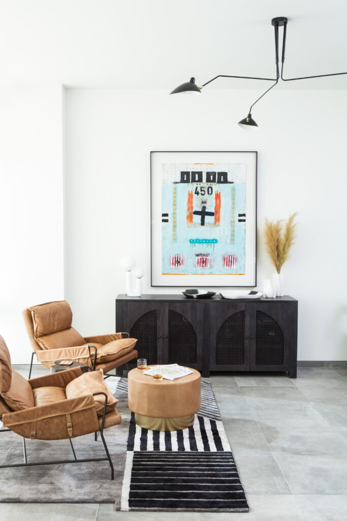 A black, gray and white graphic rug sits under two leather chairs and a leather ottoman in a living room with a black console and modern art