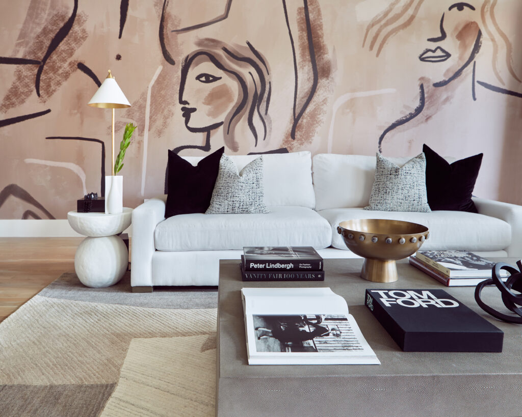 A white sofa with black and gray pillows in a living room with a pink mural covered in illustrations of female faces