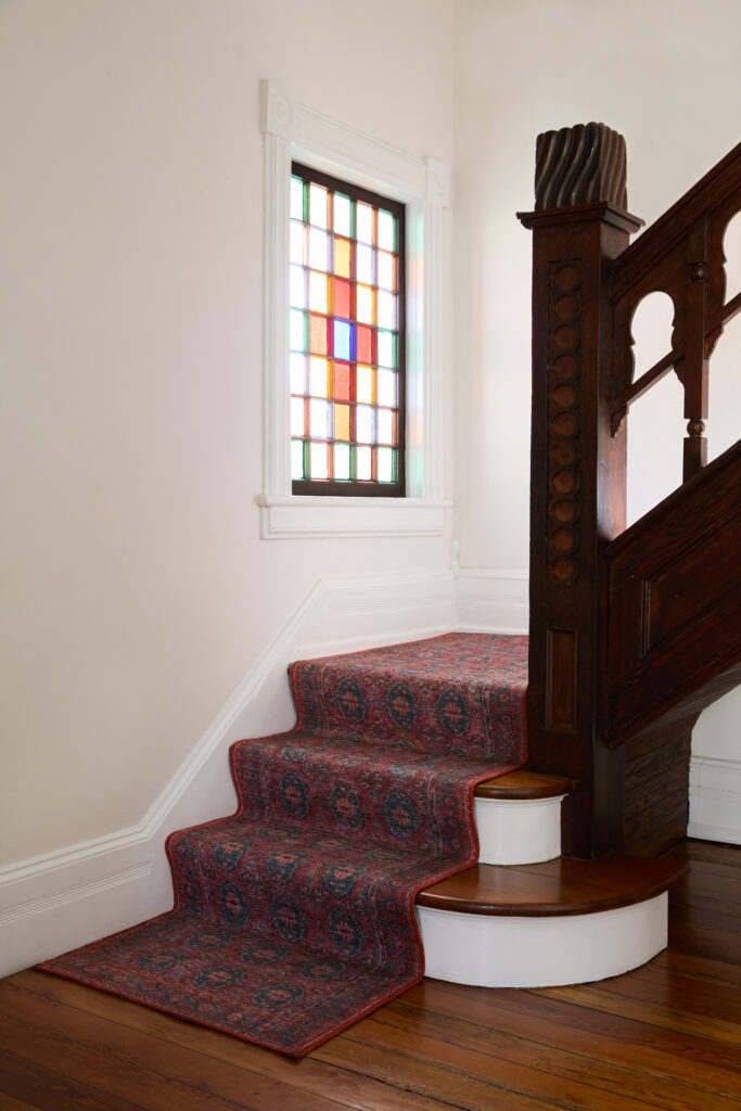A blue and red patterned stair runner goes up a staircase with dark wood banister and stained glass window