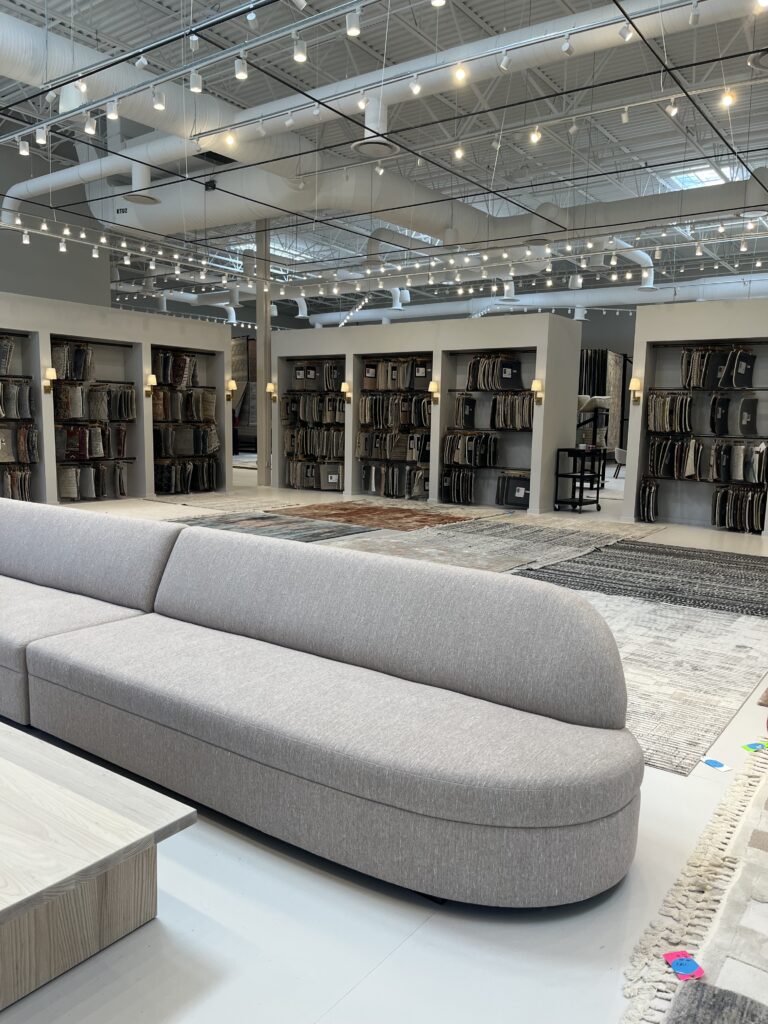 An oblong gray sofa in a white showroom with rug samples in the background