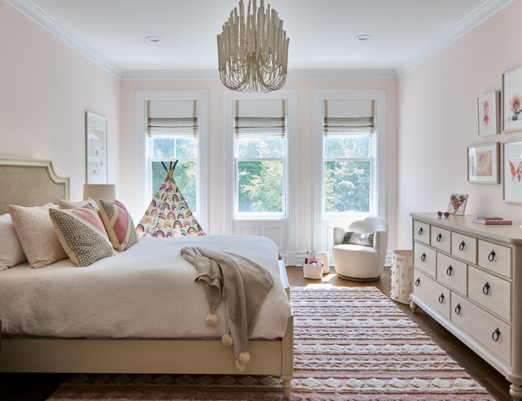 A bedroom with pink and white rug and bedding, overhead light, white walls, and white dresser